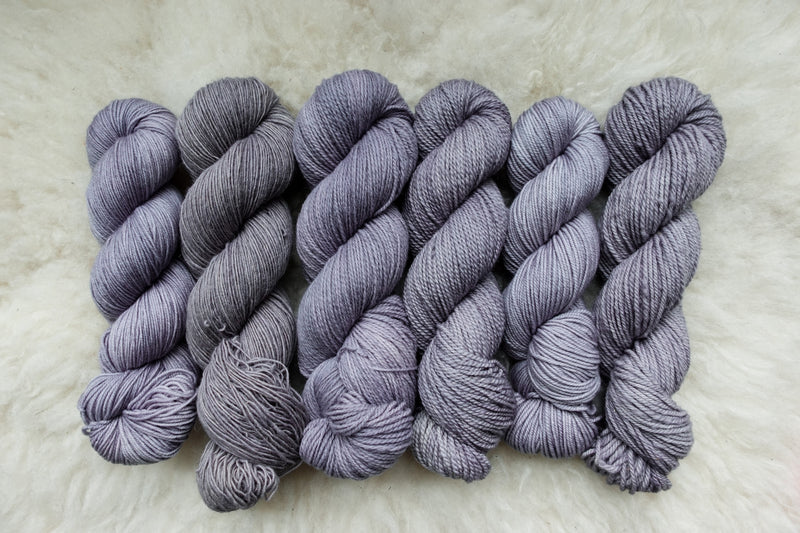 Six skeins of yarn, all different bases, have been hand dyed a light purple. They lay on a sheepskin rug.