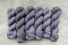 Six skeins of yarn, all different bases, have been hand dyed a light purple. They lay on a sheepskin rug.