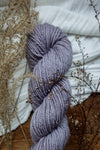 A worsted weight skein of hand dyed, light purple yarn sits on a tabletop next to dried flowers and grasses.
