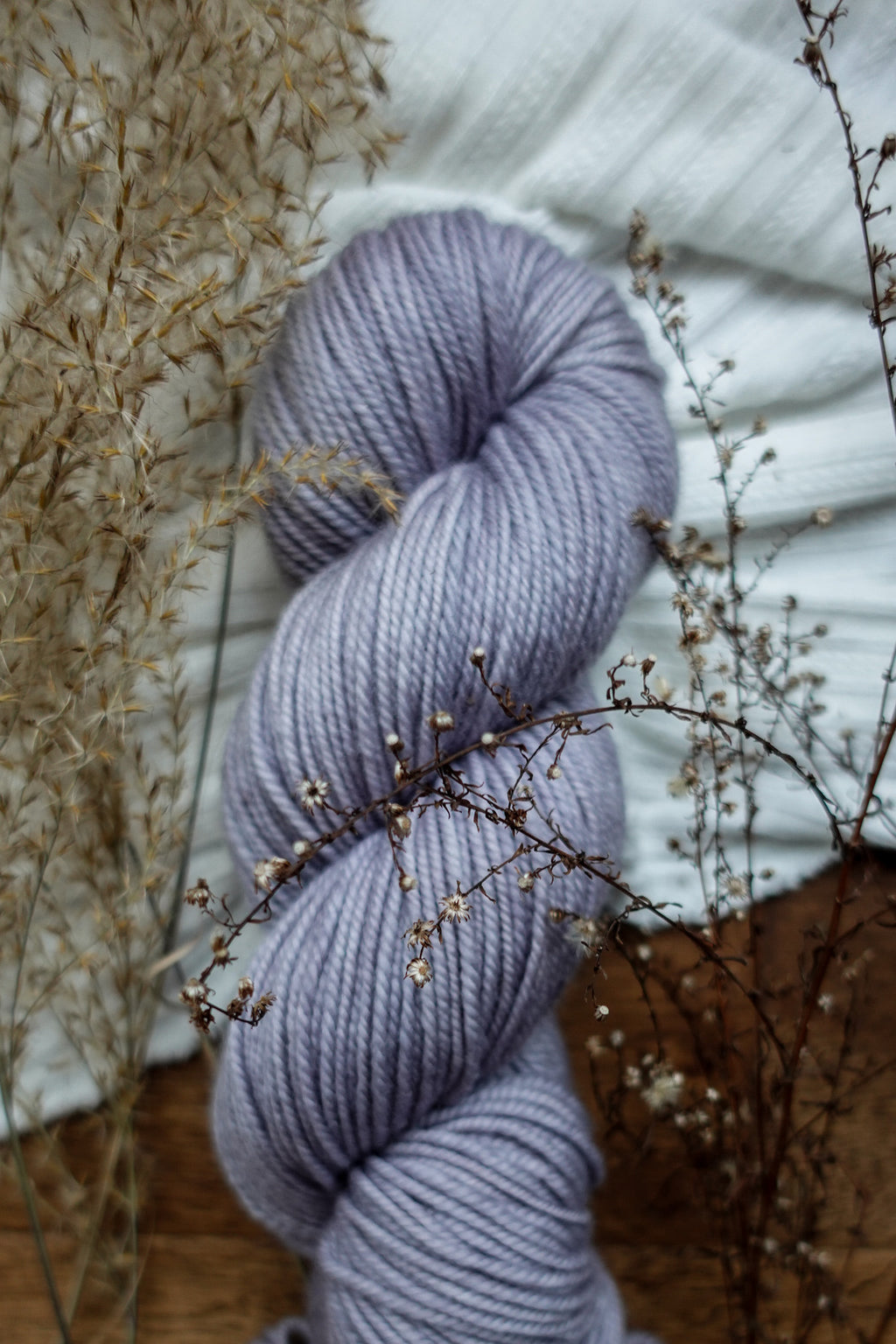 A naturally dyed skein of light purple yarn sits on a tabletop next to dried flowers.