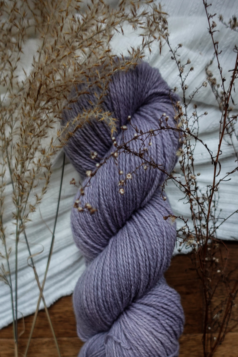 A light purple skein of hand dyed yarn sits on a tabletop next to dried flowers and grasses.