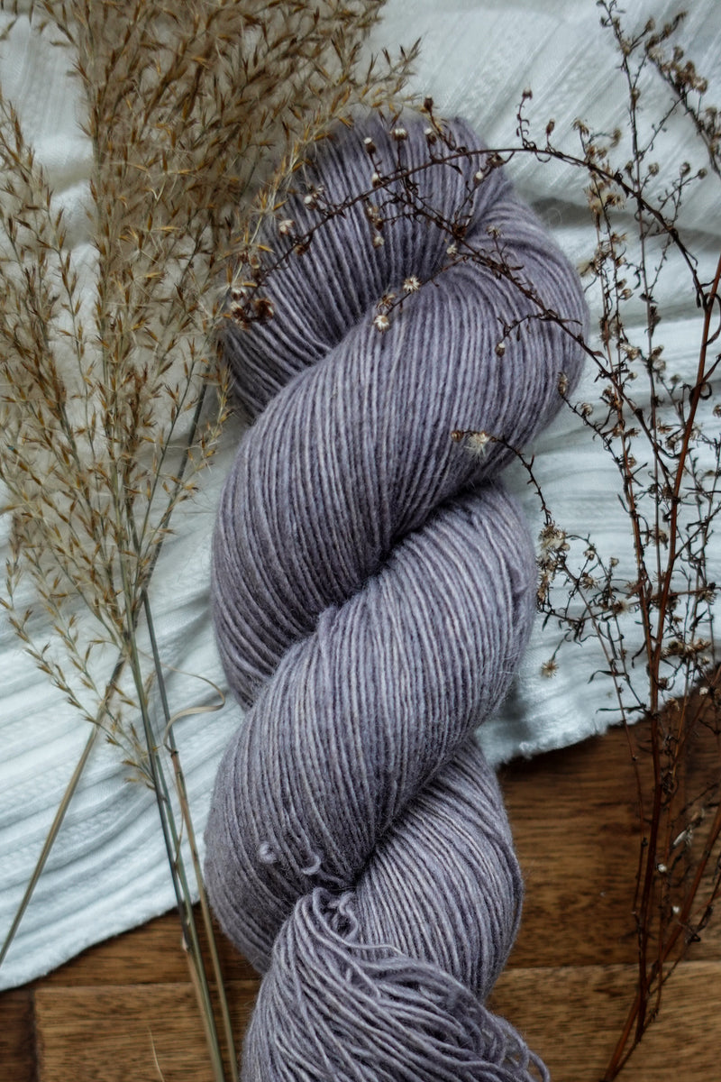 A light purple, naturally dyed skein of yarn sits on a tabletop next to dried flowers.