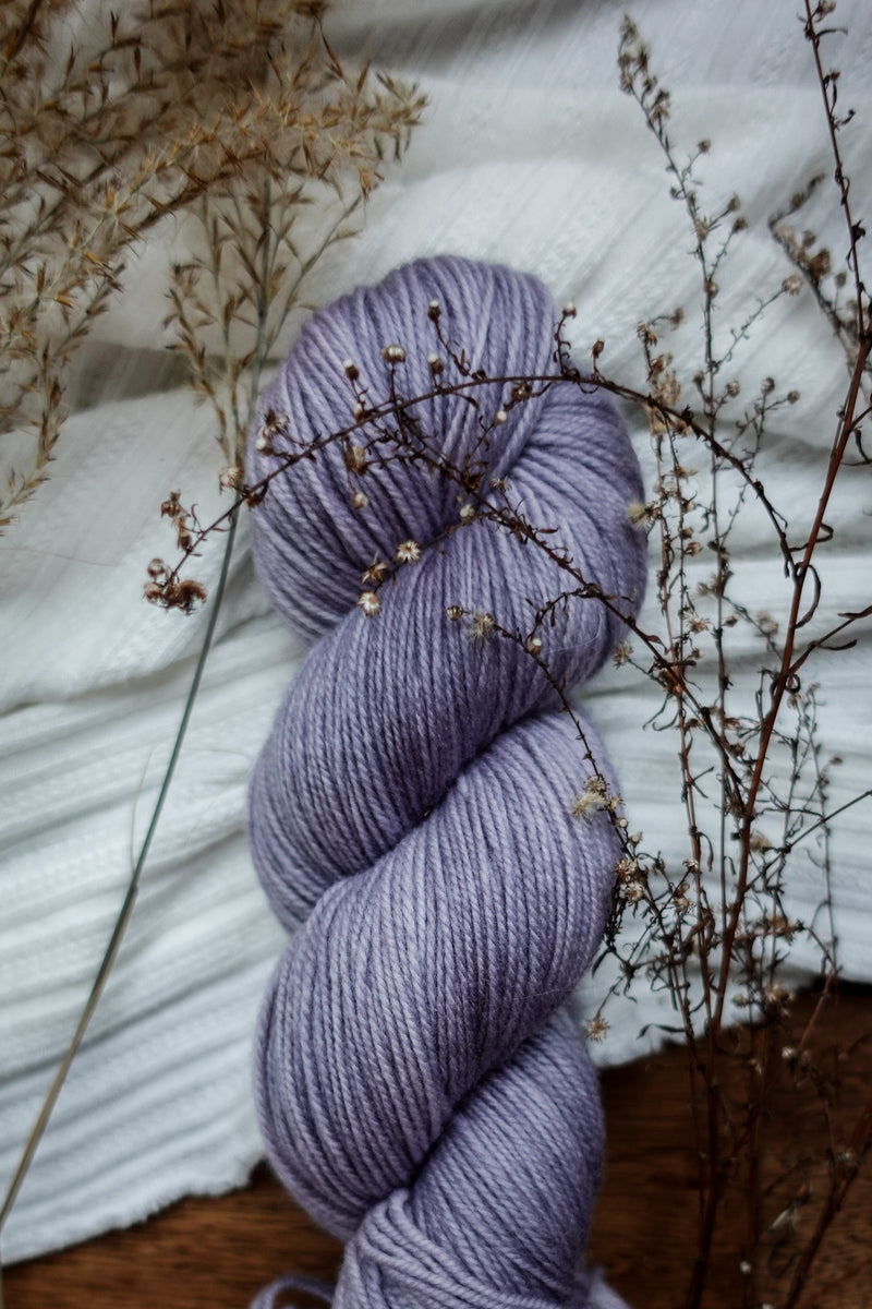 A skein of naturally dyed, light purple yarn sits on a tabletop next to dried flowers.