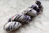 A skein of worsted weight, natural fiber yarn has been hand dyed purple and white. It lays on a wool background.