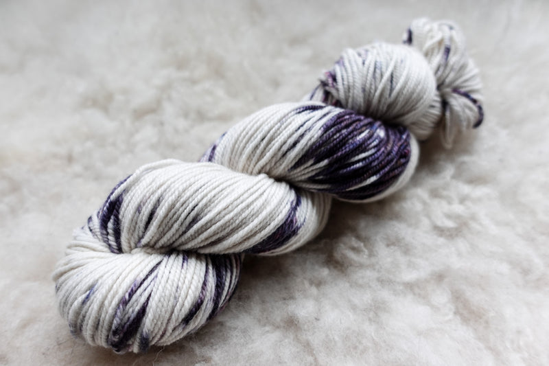 A skein of worsted weight, natural fiber yarn has been dyed in white and purple. It lays on a wool background.