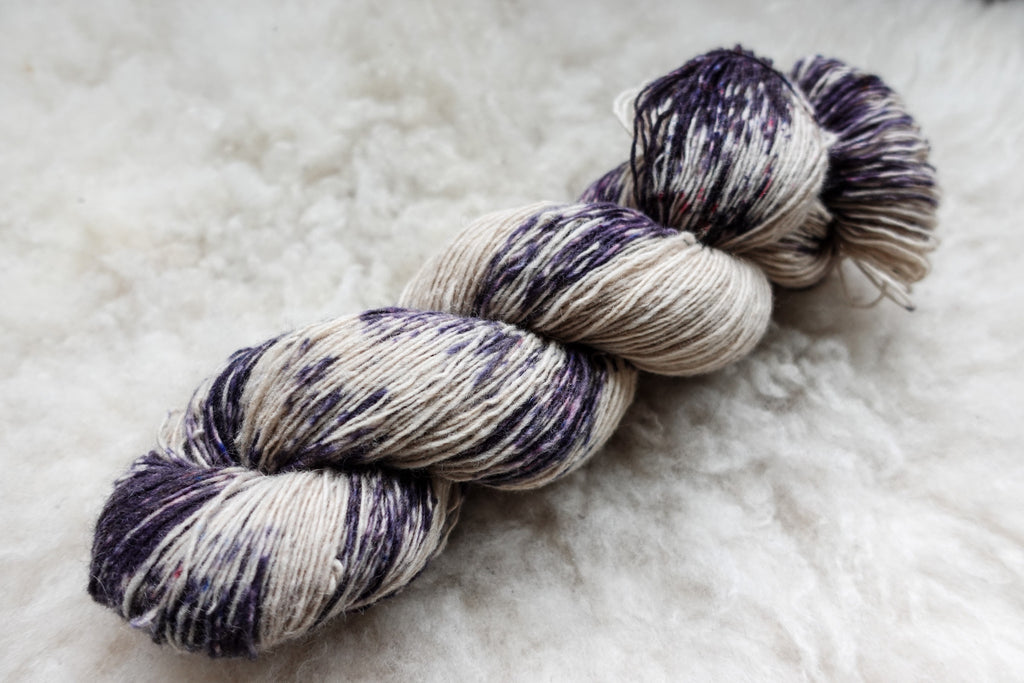 A variegated skein of yarn has been hand dyed with splashes of purple. It lays on a wool background.