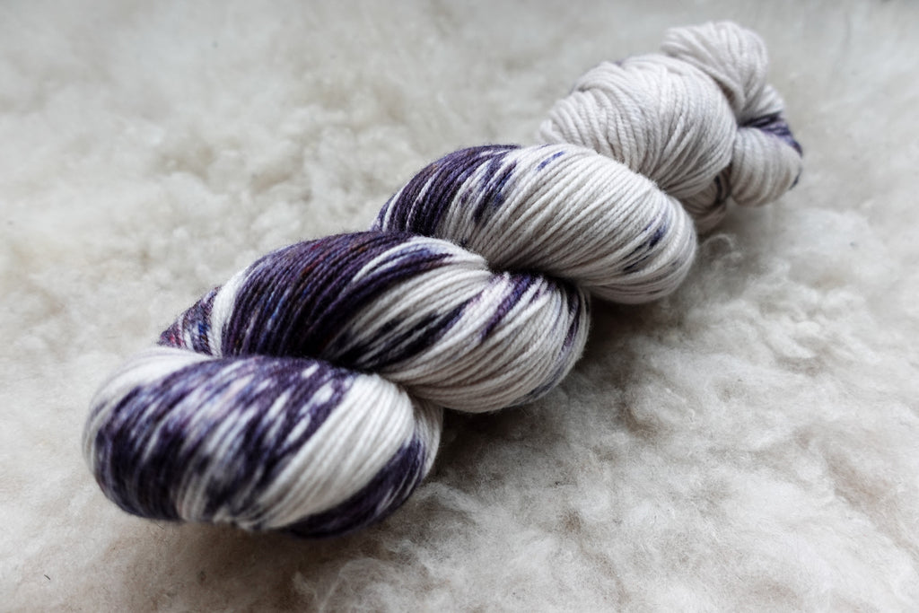 A natural fiber skein of yarn has been dyed in purple and white variegation. It lays on a wool background.