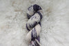 A skein of natural fiber yarn has been hand dyed with splashes of purple and white. It lays on a wool background.
