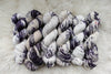 Six skeins of yarn, all different bases, have been hand dyed in a purple and white variegation. They lay on a sheepskin rug.