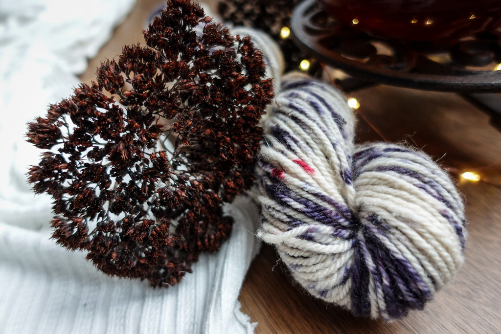 A skein of naturally dyed, purple and white yarn sits on a tabletop. Dried flowers and fairy lights can be seen in the background.