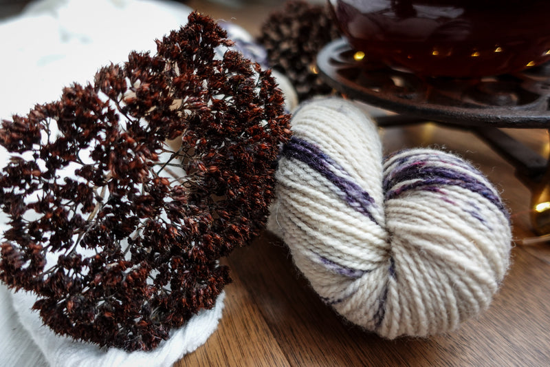 A hand dyed skein of natural fiber, purple and white yarn sits on a tabletop. Dried flowers and a teapot can be seen in the background.
