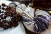 A skein of yarn is pictured close up to show the purple and white variegation. Dried flowers and a teapot can be seen in the background.