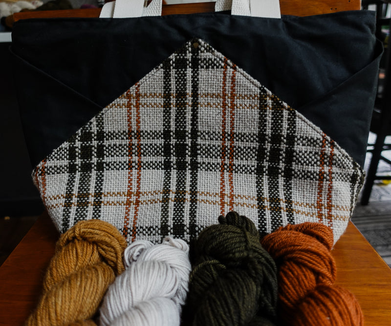 A black canvas and orange and white flannel project bag sits on a chair. Four skeins of yarn sit in front of the bag.