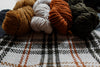 Four skeins of yarn lay on top of a project bag. The camera focuses on the orange, white, and black handwoven flannel exterior.