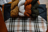 A black canvas and orange and white flannel knitting project bag lays on a chair. Four skeins of hand dyed yarn lay on top.