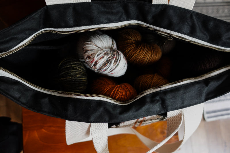 The top of a black canvas project bag is unzipped, showing six skeins of yarn inside. More space for extra skeins remains.