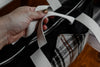 A woman's hand holds the two smaller white woven straps on a knitting project bag. There are two larger white straps. The exterior of the bag is made of black canvas and an orange and white flannel.