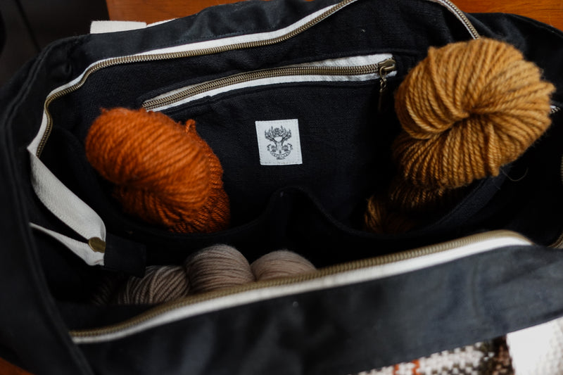 Seen from above, the top of a black canvas and orange and white flannel knitting project bag is unzipped. The black cotton lining as a zippered pocket. Three skeins of yarn are inside the bag.