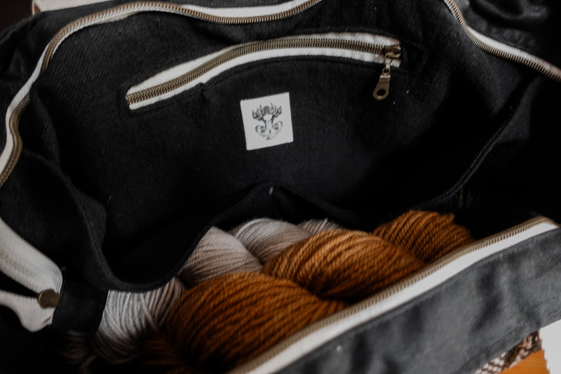 Seen from the side, the top of a black canvas and orange and white flannel knitting project bag is unzipped. The black cotton lining as a zippered pocket. Two skeins of yarn can be seen at the bottom of the bag.