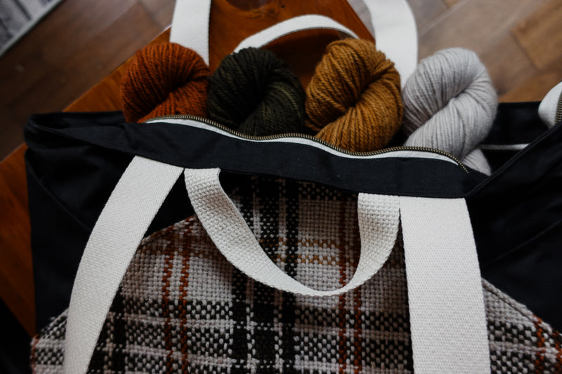A black canvas and orange and white flannel project bag lays on its. Four skeins of yarn are inside. The bag has two small handles and two longer tote handles.
