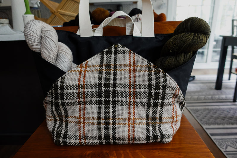 A black canvas and orange and white flannel knitting project bag sits on a chair. Two skeins of yarn are in the pockets, created by the angles of the canvas and flannel sections.