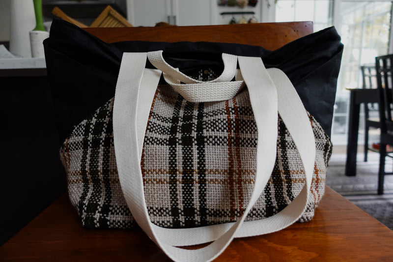 A black canvas and orange and white flannel knitting project bag sits on a chair. The camera focuses on two small white handles and the two larger white tote straps.