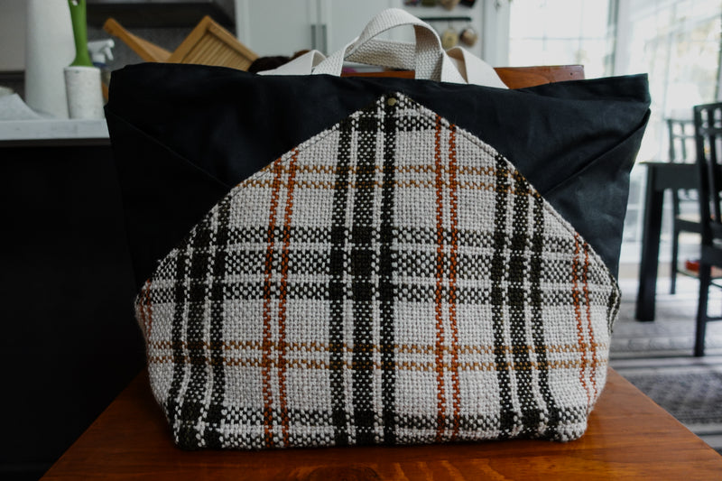 A large, tote shaped knitting bag sits on a chair. It's made of orange, white, and black woven plaid fabric and black canvas.
