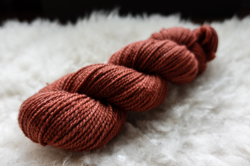 A natural skein of yarn ahs been hand dyed a brick red. It lays on a sheepskin and is seen from the side.