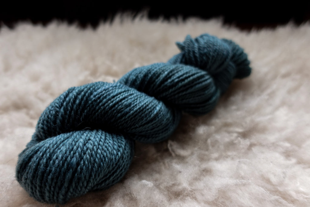 A natural skein of yarn has been hand dyed an indigo blue. It lays on a sheepskin and is seen from the side.
