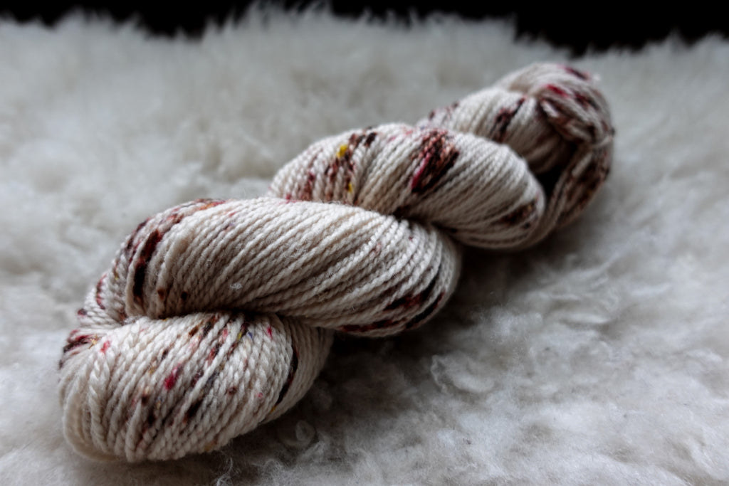 A hand dyed skein of white yarn with burgundy speckles lays on a sheepskin and is seen from the side.