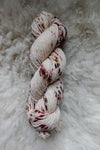 Seen from above, a white skein of yarn, naturally dyed with burgundy speckles, lays on a sheepskin.