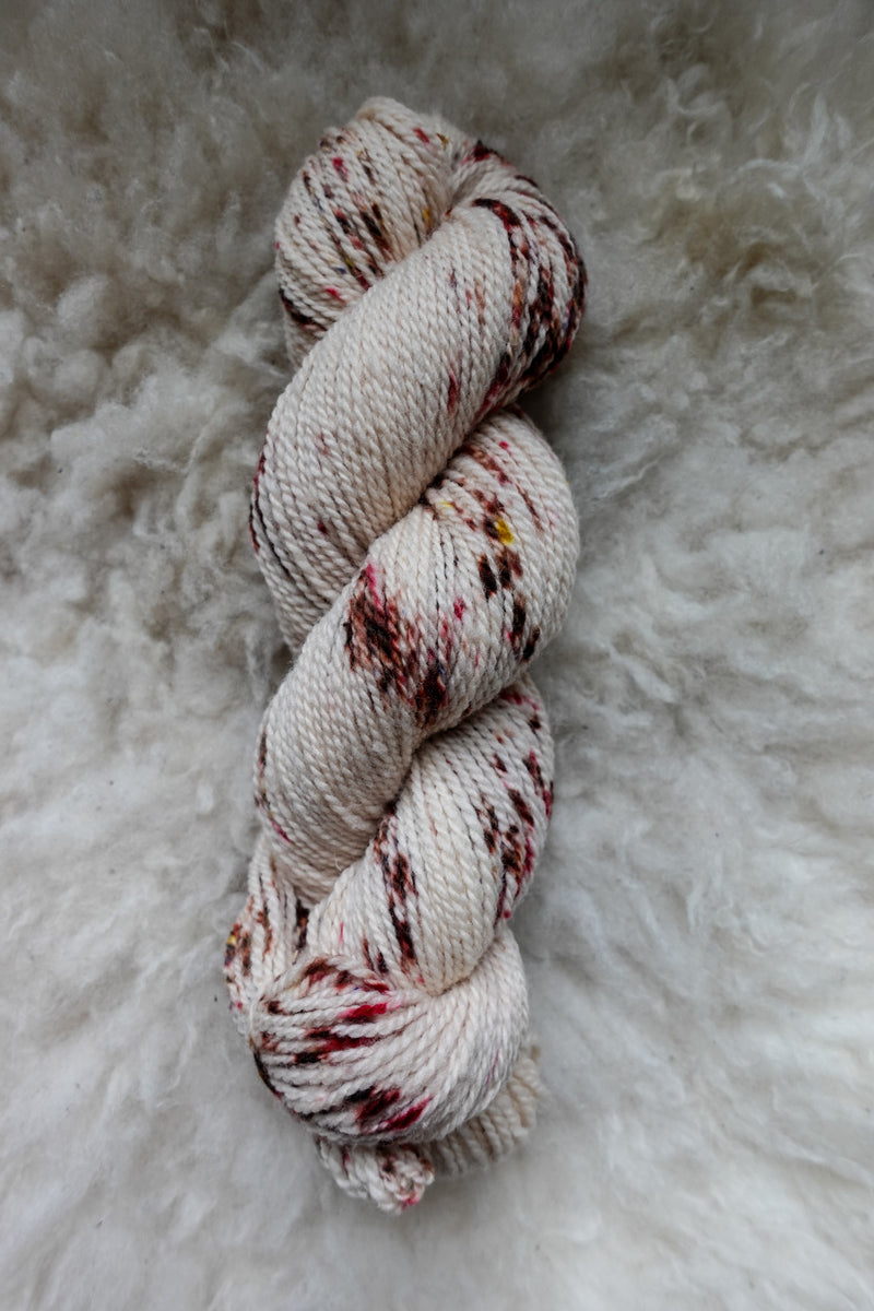 Seen from above, a white skein of yarn, naturally dyed with burgundy speckles, lays on a sheepskin.