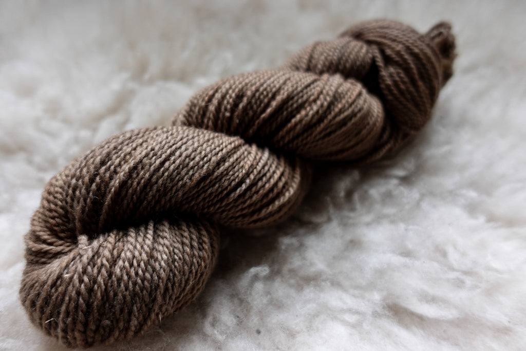 A natural skein of yarn has been hand dyed a light brown. IT lays on a sheepskin and is seen from the side.
