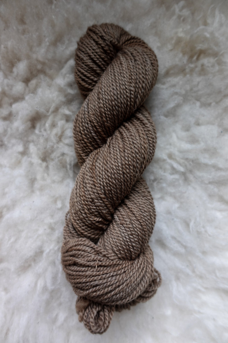 Seen from above, a light brown skein of hand dyed yarn lays on a sheepskin.