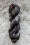 Seen from above, a brown, white, and grey variegated skein of hand dyed yarn lays on a sheepskin.