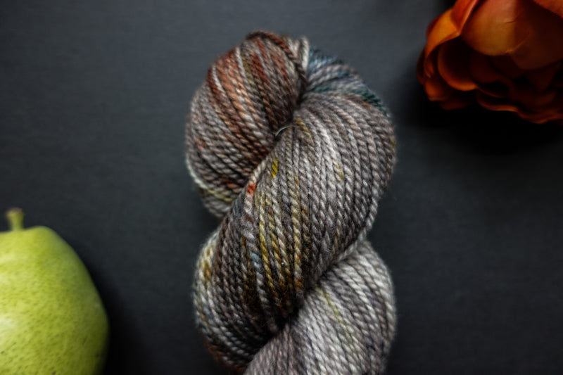 Seen close up, a variegated brown, grey, and white skein of naturally dyed yarn lays on a black surface next to an orange-red flower and a pear.