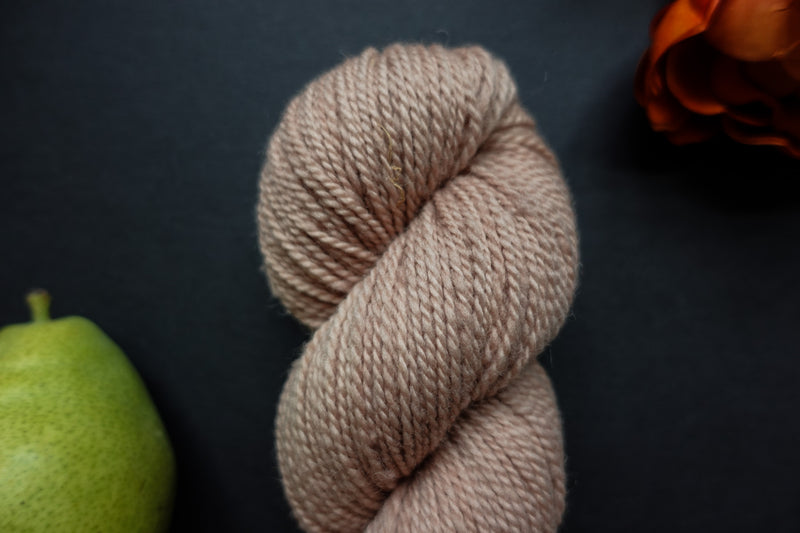 Seen close up, a hand dyed skein of light pink-beige yarn lays on a black surface next to an orange-red flower and a pear.