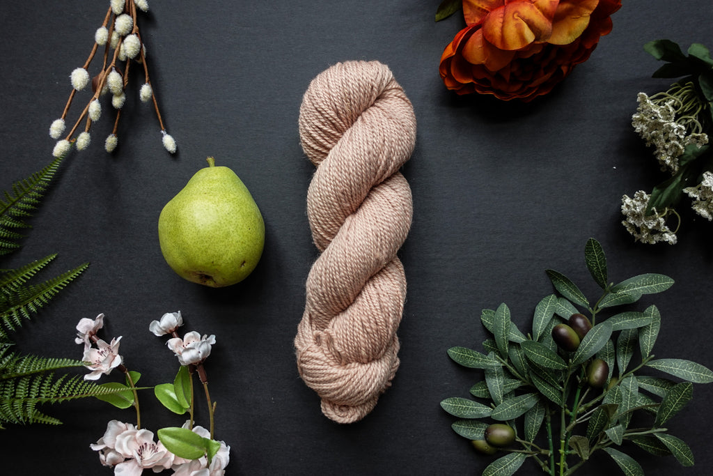 A skein of light beige DK weight yarn lies on a black surface. It's surrounded by flowers, branches, an orange rose, and a pear.