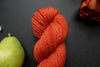 Seen close up, an orange-red skein of naturally dyed yarn lays on a black surface next to a matching flower and a pear.
