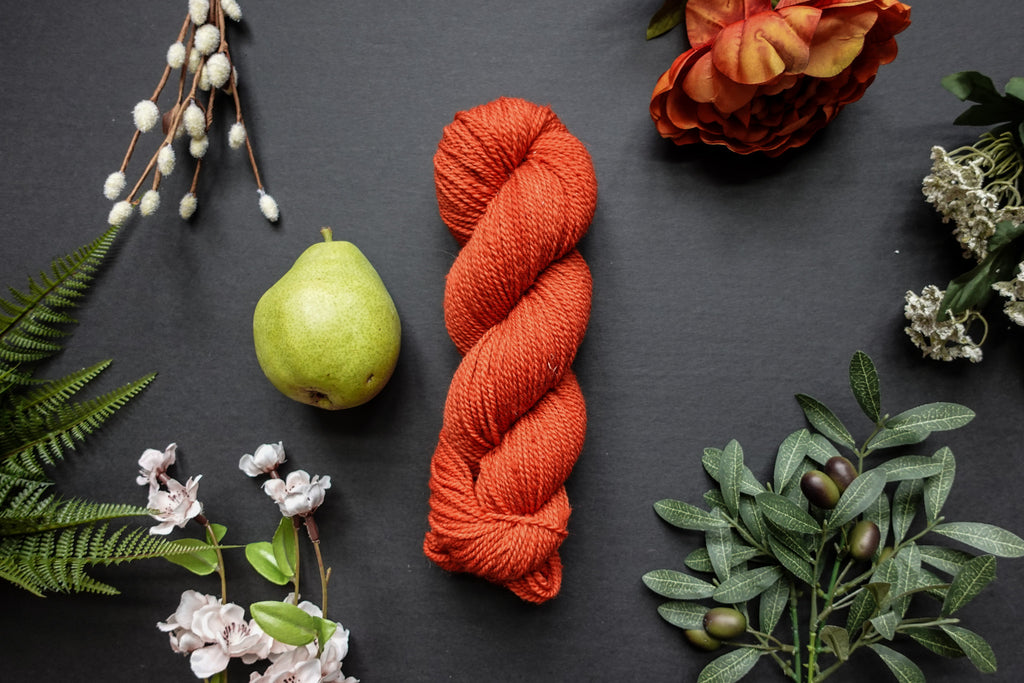 A orange-red skein of DK weight yarn lies on a black surface. It's surrounded by flowers, branches, an orange rose, and a pear.