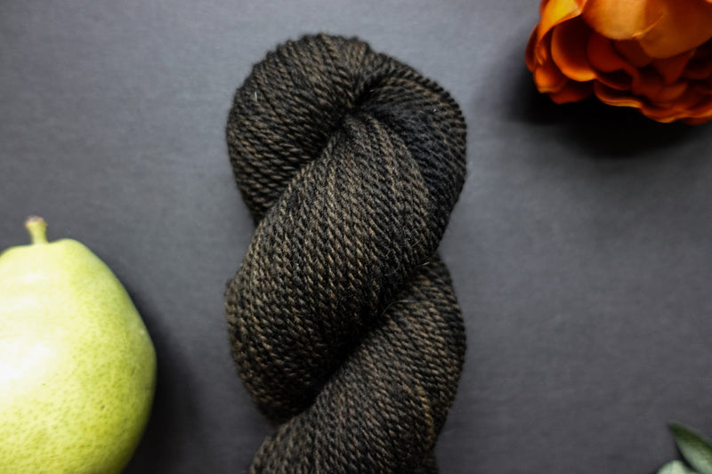 Seen close up, a muddy green, almost black, skein of hand dyed yarn lays on a black surface. It is surrounded by an orange-red flower and a pear.