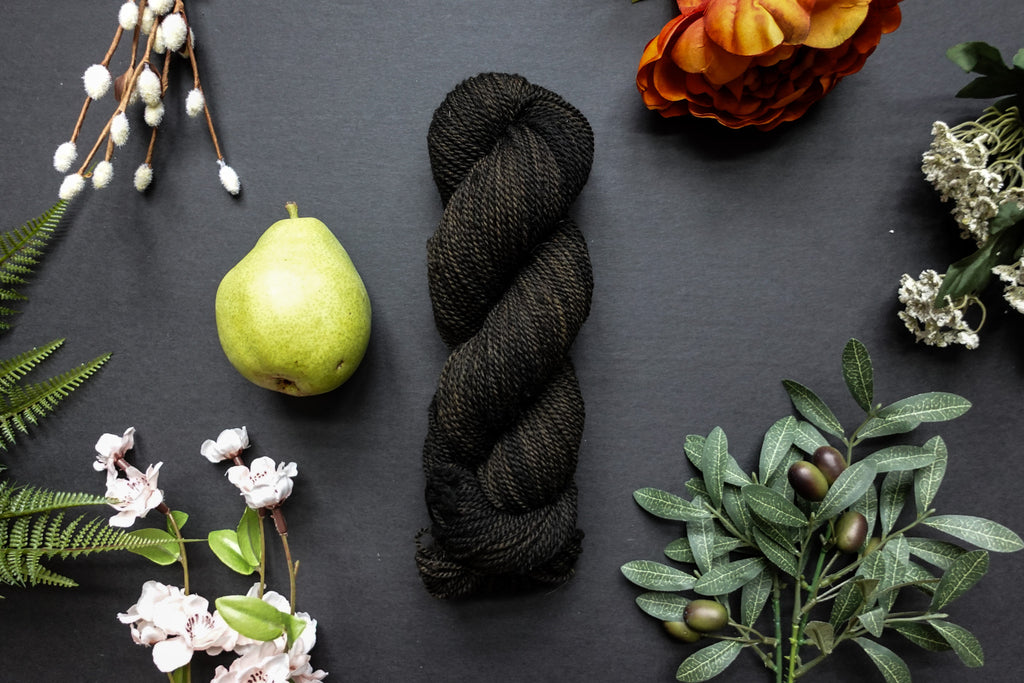 A dark, muddy green, almost black, skein of DK weight yarn lies on a black surface. It's surrounded by flowers, branches, an orange rose, and a pear.