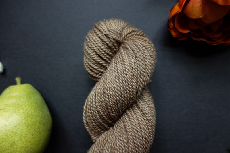Seen up close, a light brown skein of hand dyed yarn lays on a black surface next to an orange-red flower and a pear.