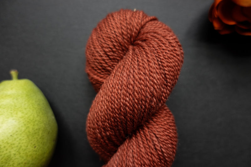 Seen close up, a brick red skein of hand dyed yarn lays on a black surface next to a per and an orange-red flower.