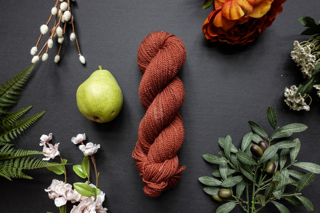 A brick red skein of DK weight yarn lies on a black surface. It's surrounded by flowers, branches, an orange rose, and a pear.