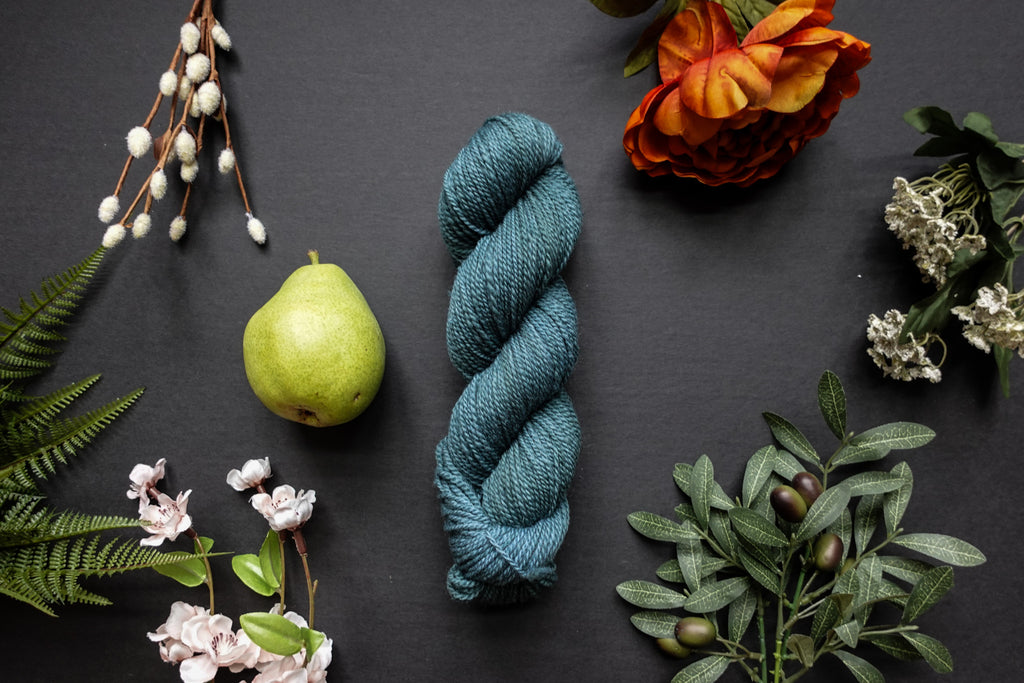 A skein of blue sport weight yarn lies on a black surface. It's surrounded by flowers, branches, an orange rose, and a pear.