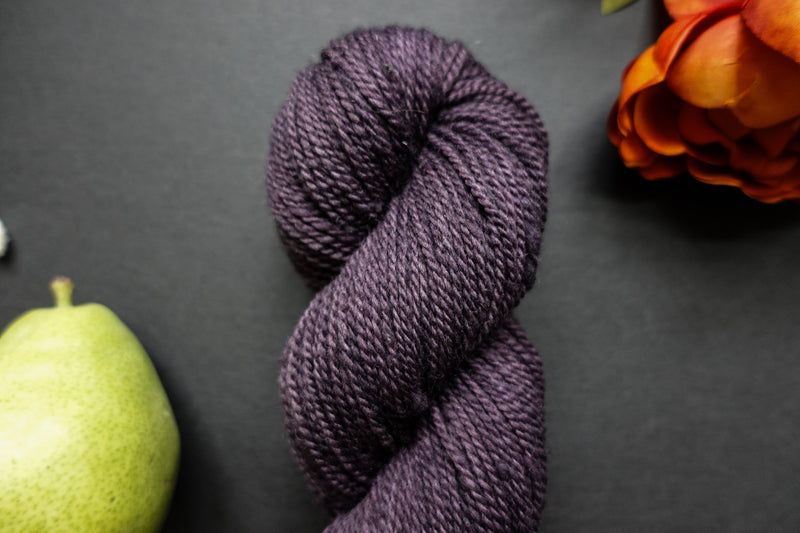 Seen close up, a deep purple skein of naturally dyed yarn lays on a black surface next to an orange-red flower and a pear.