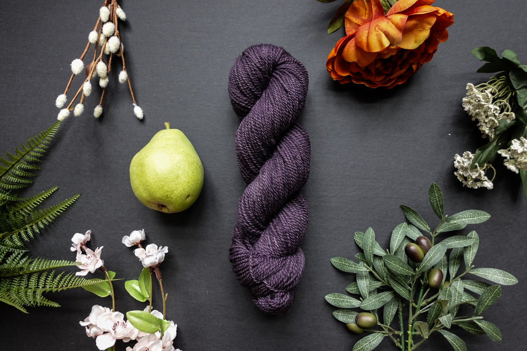 A skein of deep purple DK weight yarn lies on a black surface. It's surrounded by flowers, branches, an orange rose, and a pear.