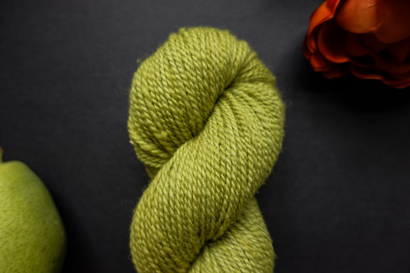 Seen close up, a bright green skein of naturally dyed yarn lays on a black surface next to an orange-red flower and a pear.