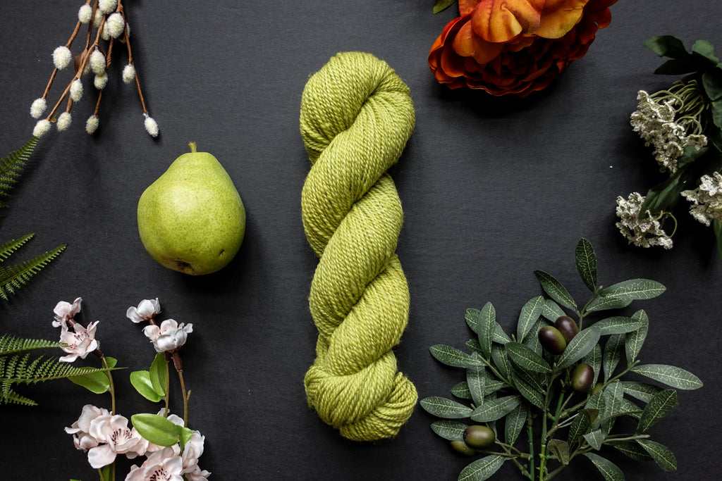 A bright green skein of DK weight yarn lies on a black surface. It's surrounded by flowers, branches, an orange rose, and a pear.